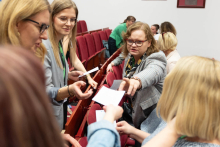 Grupa osób w sali konferencyjnej z czerwonymi krzesłami. Część osób stoi, część siedzi, przekazując sobie kartki papieru. W tle widoczne są inne osoby oraz fragmenty ścian z ramkami.”