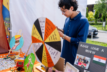 A young man is standing at a handicraft stand. He has dark hair and a blue shirt. In the foreground is a colorful roulette table with English inscriptions, including 'Pick with your eyes closed.' Next to her are decorative boxes, pictures and a board with the Indian flag. 