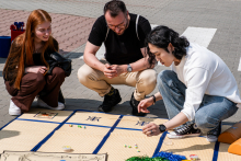 Three people sit on a mat spread out on the sidewalk and participate in a board game. The mat is divided into squares and contains pawns and cards.