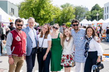 Seven people stand outside during the event. They are the dean's authorities and representatives of the student government. background of exhibition tents and trees. The people are dressed variously: including a man in a red jacket, a woman in a white blouse and green dress, a woman in a flowery skirt, and a woman with a scarf around her neck.