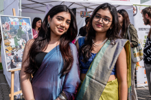 Two young women in traditional costumes stand side by side. One has a blue and purple sari, the other a green and yellow sari with a blue blouse. Both have long dark loose hair. In the background are booths with posters and banners, including the words 'Pakistan' and 'Groceries and more from Pakistan'.