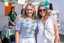 Two young women with green garlands around their necks stand in front of a stand with Irish flags, balloons and a landscape poster. One is wearing a blue and white striped blouse and white pants, while the other is wearing a white sleeveless blouse with a light-colored handbag. Both have light-colored hair. They are smiling for the photo.