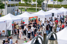 A row of white tents around which people gather. Participants talk and take part in various activities. Some tents decorated with balloons and banners. In the background are trees and buildings.