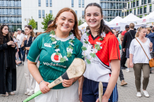 Two young women are standing side by side, holding hurling sticks. One is wearing a green Vodafone jersey and white shorts, the other a red jersey with white elements and blue shorts. Both have floral garlands around their necks. In the background are people and tents.