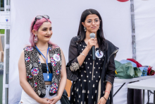 Two young women stand side by side against the backdrop of a white tent. One has pink hair, a colorful sequined vest and a white top, while the other is wearing a black dress with a white pattern and holding a microphone to which she is speaking. A white tent can be seen in the background.