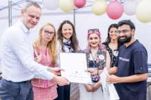 Six people stand under a tent outside. The three people on the left hold together a diploma, which they hand to the person on the right. The faces have been blurred. In the background are balloons in white, pink and yellow.