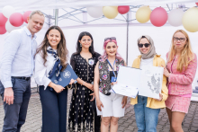 Six people stand under a white tent on a paved surface. In the background are balloons in red, white and yellow. One person holds an open briefcase with documents and a diploma. The people's outfits range from elegant to casual. 