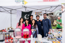 A group of people - young women and young men stand under a white tent with the flag of Pakistan. They are smiling for a photo. On a table in front of them are two dispensers of pink drinks, bottles of sauces, books and other items. 