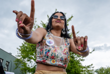 A young woman with dark loose hair a top with a floral pattern and a transparent panel on her neckline raises her hands, pointing her fingers upward. On her wrists are bracelets, in her ears earrings. In the background are trees and a section of a building.