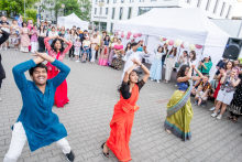 Young women and young men in traditional costumes dance on a paved square in front of a modern building. Faces are covered. In the background is a crowd of people, some taking pictures or recording. White tents and balloons in pink, white and gold are visible.