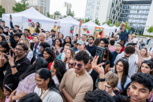 A crowd of people gathered outside in front of white exhibition tents. In the background, modern buildings. Some people are taking photos or videos with their phones. 
