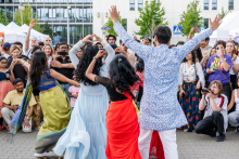 A group of people in colorful traditional costumes dance on a cobblestone surface during an outdoor event. In the background is a crowd of observers taking photos, tents and office buildings. The scene takes place outside.