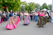 Three women are dancing outdoors in colorful traditional costumes. One has a pink skirt and a red fan, another a purple skirt with a red sash, the third a black skirt and a pink blouse with a blue fan. In the background are spectators, trees and a tent.