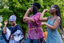 Three people are dancing in the open air. One has a traditional African outfit and a headscarf on her head, another has a pink blouse with a pattern and a black cap, and the third has a blue dress with white dots. In the background are trees, cars and other people.
