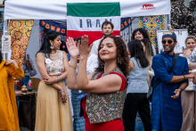 A group of people stand in front of a stand with the word “IRAN” on the background of the Iranian flag. In the foreground, a woman in a red blouse and vest claps her hands. Some people dressed in traditional costumes. In the background are colorful decorations and participants in the event.
