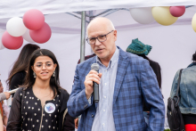 A man - the rector of WUM - in a blue checkered jacket holds a microphone and speaks. Next to him stands a young woman with long dark hair in a black dress with white buttons. In the background are balloons in white, pink and yellow and participants in the event.