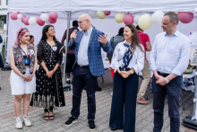 Five people stand under a white tent outside. The man in the center - the rector of WUM - holds a microphone and speaks. In the background are balloons in white, pink and yellow and other participants in the event. The ground is paved.
