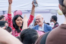 A group of people outside during an event. A woman in a red dress with patterns lifts her phone while taking a photo or video. Henna is visible on her hands. Next to her stands a man in a blue jacket. In the background are colorful balloons and event participants.