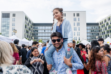 A crowd of people outside during a festival. A young man in a shirt holds a girl in a blue sweatshirt and pink pants on his shoulders. In the background are modern buildings and white exhibition tents, with people around watching the booths and talking.