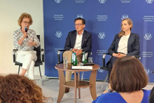 Three speakers sit on stage in front of a blue backdrop with the logo of the Warsaw Medical University. One of them is holding a microphone. A table with water in front of them. In the background, visible participants sitting with their backs to the camera.