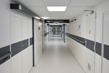 A bright hospital corridor with white walls and gray stripes. At the end, a glass window. On the left, a door with a sign reading “HEMODYNAMICS PRACTICE I.” Rectangular lamps on the ceiling.