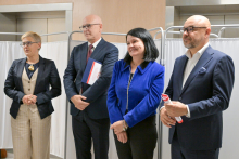Four people stand side by side against a background of white screens. The first person in a light suit and dark jacket. The second in a dark suit, holding documents. The third in a blue jacket. The fourth in a dark suit, holds a folded document.