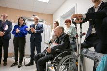 A group of people in elegant clothes stand in a bright room with white walls. In the background are white medical screens. In the foreground, a man in a wheelchair holds crutches. Next to him stands another man, leaning on crutches.