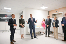 Six people stand in a bright room. Three on the left are dressed formally, two are holding documents. In the middle, a man in a suit gestures. On the right three people, one with a briefcase. In the background is a table with drinks and snacks and an information board about the invasive cardiology department.