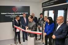 A group of people stands in front of the entrance to the Invasive Cardiology Unit. A woman in a gray jacket cuts a red ribbon held by two men. On the left is a black board with white lettering about the labs and patient rooms. On the right, a door with a similar sign.