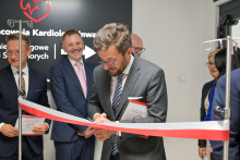 A man - Prof. Pawel Wlodarski, dean of the Faculty of Medicine - in a suit cuts the red-and-white ribbon with scissors. In the background, people in formal attire. Behind them is a black banner with white lettering “Invasive Cardiology Laboratory” and a red heart with an EKG line.