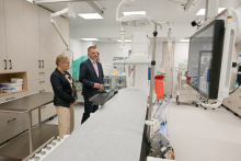 Two formally dressed people stand in a modern medical office. One is holding documents. To the right is a large monitor, to the left medical equipment and cabinets. The dominant colors are white and gray.