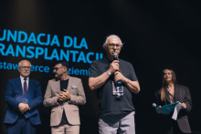 Four people are standing on the stage: three men and one woman. The man on the left is wearing a blue suit and holding a statuette. Next to him is a man in a beige suit. Next is an older man in a black T-shirt holding a microphone. The woman on the right in a dark outfit holds a microphone and briefcase. A sign in the background reads “FOUNDATION FOR TRANSPLANTATION.”