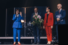Four people are standing on the stage. The woman on the left is wearing a blue suit and red shoes, holding a microphone. Next to her is a man in a dark suit with a bouquet of flowers. Next is a woman in a red suit. On the right, a man in a dark suit with a briefcase with the university logo. In the background, drums and instruments behind transparent covers.