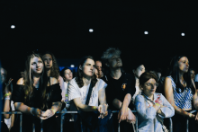 GGrupa osób stoi za metalowym ogrodzeniem podczas koncertu. W tle ciemne niebo i jasne światła sceniczne. Większość patrzy w stronę sceny, niektórzy opierają ręce na barierce. Ubrania różnorodne – koszulki, bluzy. Twarze zamazane ze względów prywatności.