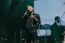A man in a black shirt with white patterns sings on stage. He holds a microphone in his right hand. In the background, a large screen shows an enlarged image of him. The stage is illuminated by green spotlights.