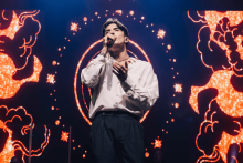 A man in a white shirt and dark pants stands on stage with a microphone to his lips. He looks as if he is singing. The background is colorful, with orange and red patterns resembling clouds and stars. In the center of the background, a circular pattern surrounded by bright dots is visible.