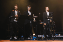 Three musicians in black suits and white shirts stand on stage. They play trumpet, saxophone and trombone. In front of them are microphones on tripods and music stands with sheet music. On the floor, water bottles and electronic equipment. The background is dark, illuminated by warm, golden spotlights.