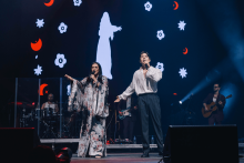 Two singers are singing on stage. The woman on the left is wearing a long, patterned kimono dress and holding a microphone. The man on the right in a white shirt and dark pants, also holding a microphone. In the background is a screen with animation: a white silhouette of a woman, red crescents and white flowers on a black background. Musicians with instruments can also be seen.