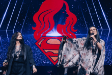 Two people are standing on the stage. On the left, a person in a dark outfit with a necklace holds a microphone. On the right, a person in a light-colored kimono with a floral pattern gestures with his hand. In the background is a red Superman symbol and the profile of a woman with long hair against a starry sky.
