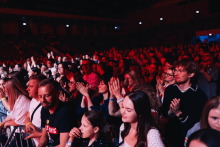 A crowd of people at a concert. Many people holding phones, recording or taking pictures. The stage illuminated with red and blue lights. People stand close together, dressed in light and dark outfits. In the background is a dark hall with spot lights.