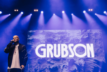 On the concert stage, illuminated by blue spotlights, stands a person in a dark jacket and white T-shirt, holding a microphone. In the background is a large screen with the words “GRUBSON” against a background of abstract patterns.