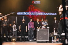 A group of people stands on a stage. In the background is a large screen with the words “ARTISTS FOR TRANSPLANTATION” and “12 HOURS FOR LIFE” and a logo. The stage is lit with warm light. Participants are dressed in a variety of attire - from everyday clothes to formal attire. To the right, public address equipment is visible.