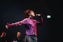A person in a purple Adidas jacket sings on stage, holding a microphone in his right hand and with his arms spread out. In the background are two musicians, one playing keyboards. The stage is dark, lit with spot lights.