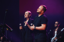 Two singers are singing on stage. The woman on the left in dark clothing holds a microphone in her right hand. The man on the right in a black T-shirt also holds a microphone. In the background is a guitarist with a white guitar and a tripod with sheet music. The stage is illuminated with purple light.