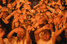 A crowd of people at a concert with their hands raised. Some are holding phones. Participants have identification bands on their wrists and different clothes. Faces are blurred. The background is blurred, the lighting in a warm orange hue.