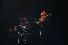 Two musicians play brass instruments. On the left is a trumpeter, on the right a trombonist. Both are dressed in dark costumes. They stand on a stage with a dark background. The trombonist's face is obscured by a square.
