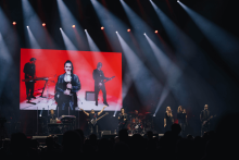A musical band performs on stage. The musicians play guitars, keyboards and drums. The vocalist stands at the front. In the background, a large screen with a red background shows a picture of the band members. The stage is illuminated by bright spotlights pointing downward.