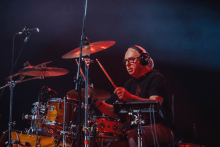The man plays a wooden drum with metal parts. He is wearing a black T-shirt and headphones. He is located on the stage. Dark stage lighting is visible in the background.
