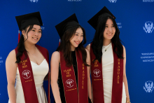 Three women in white gowns and black birettas stand in front of a blue wall with the inscription “Warsaw Medical University”. They have maroon sashes with the inscriptions “CLASS OF 2025” and “GRADUATE”.