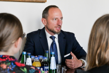 Mężczyzna w garniturze siedzi przy stole z butelkami napojów. Po bokach widoczne są sylwetki dwóch osób, uchwycone od tył